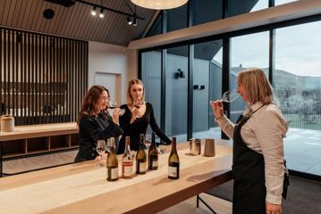 Two customers and a worker try wine in The Runholder. The floor, walls, and high-ceiling roof are a light wood with black accents in the lights and fixtures.