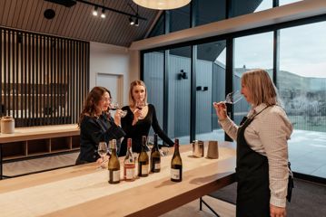 Two customers and a worker try wine in The Runholder. The floor, walls, and high-ceiling roof are a light wood with black accents in the lights and fixtures.
