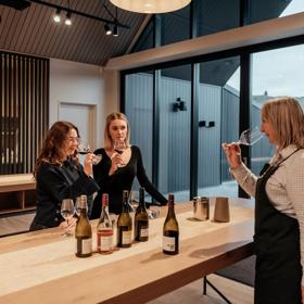 Two customers and a worker try wine in The Runholder. The floor, walls, and high-ceiling roof are a light wood with black accents in the lights and fixtures.