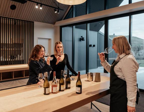 Two customers and a worker try wine in The Runholder. The floor, walls, and high-ceiling roof are a light wood with black accents in the lights and fixtures.
