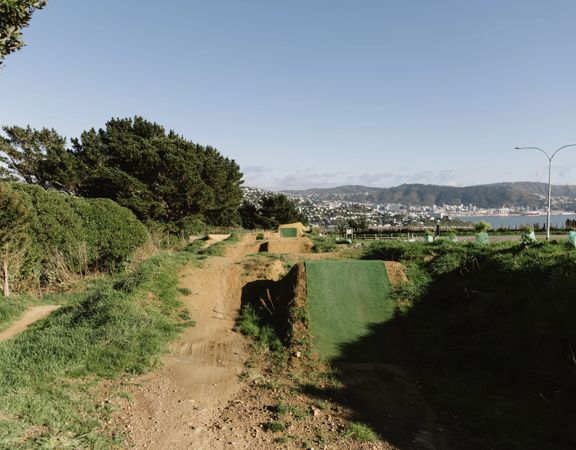 Dirt mountain bike jump tracks lined with synthetic grass, and Wellington city in the background.