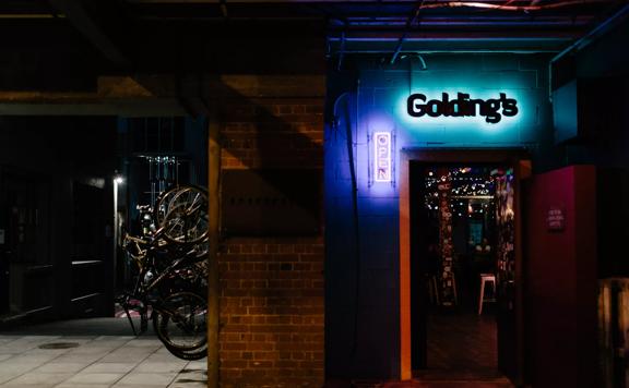 The exterior of Golding's. A door sits open under a glowing neon blue sign. To the left is Hannah's Laneway with bikes hanging on the wall.