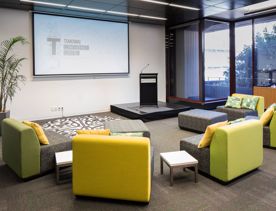 The yellow and gray couches facing a podium and projector inside Programme Rooms Te Wehenga & Malaga Pasifika, at Tiakiwai Conference Centre - National Library of New Zealand.