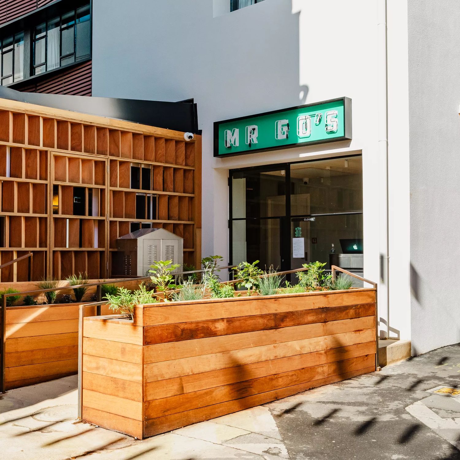 Entrance of Mr Go's restaurant with a green sign, glass doors, white walls and wooden planters leading up to the door.
