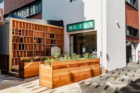 Entrance of Mr Go's restaurant with a green sign, glass doors, white walls and wooden planters leading up to the door.