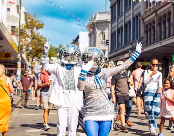 Two street performers dresses in silver and blue with disco balls on their heads are dancing and waving as a part of the CubaDupa street festival in Wellington.