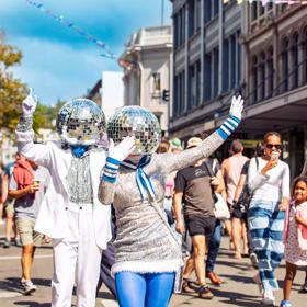 Two street performers dresses in silver and blue with disco balls on their heads are dancing and waving as a part of the CubaDupa street festival in Wellington. 