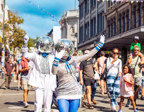 Two street performers dresses in silver and blue with disco balls on their heads are dancing and waving as a part of the CubaDupa street festival in Wellington. 