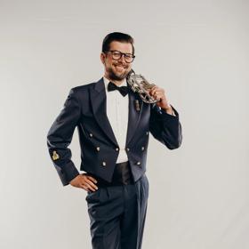 A person in a navy blue tuxedo stands with their left leg crossed over the right, their left hand on their hip, and their right hand holding a trumpet over their right shoulder. 