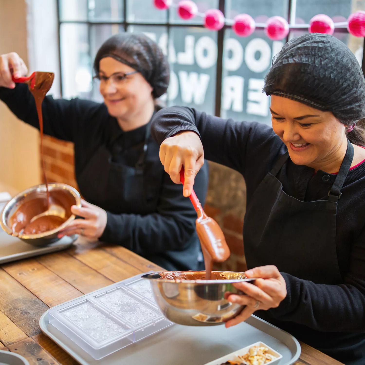 Two people are mixing melted chocolate in silver bowls at Wellington Chocolate Factory located in Te Aro, Wellington.