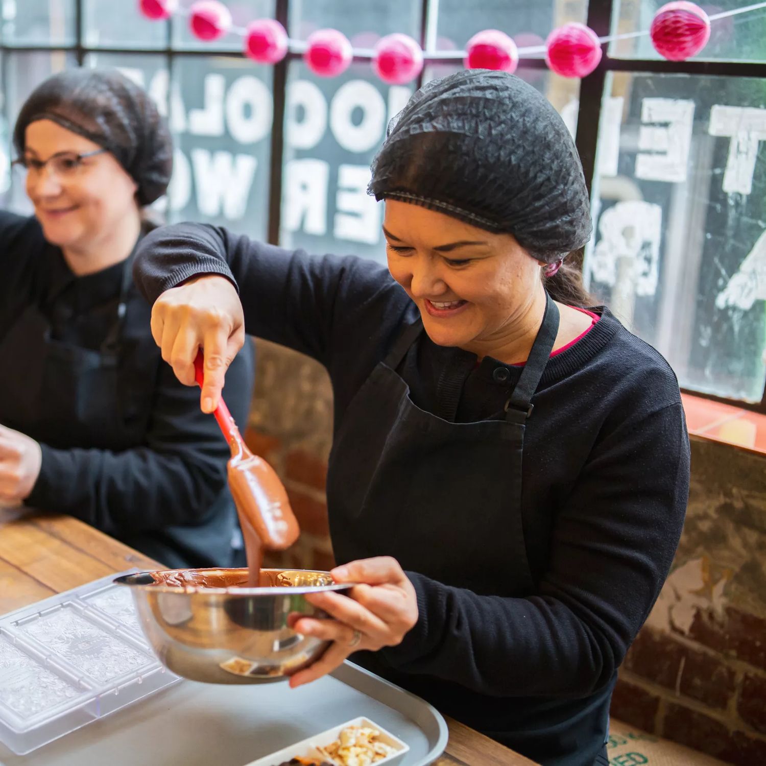 Two people are mixing melted chocolate in silver bowls at Wellington Chocolate Factory located in Te Aro, Wellington.