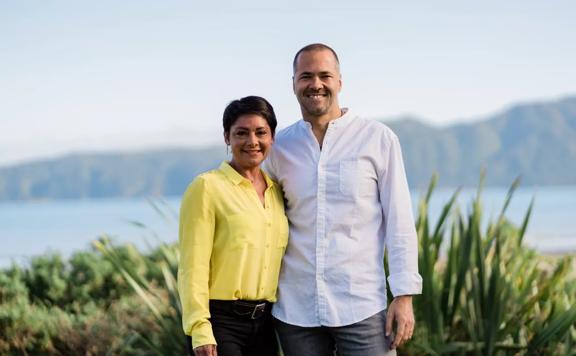 Val Reitnauer, has short brown hair wearing a yellow blouse and black pants, stands with her husband Timo Reitnauer, in a white shirt and grey jeans, with water and hills in the background.