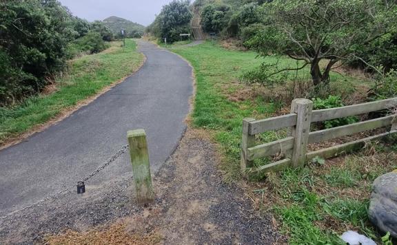 Trail entrance showing a one metre wide gap in a fence leading towards a driveway. The surface is a mix of gravel, concrete and grass.