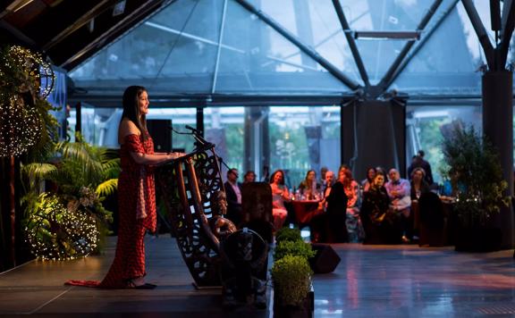 Person on stage speaking into a podium microphone out to many people in the background seated at tables. Dim mood lighting surrounds the area with spotlight on the speaker.