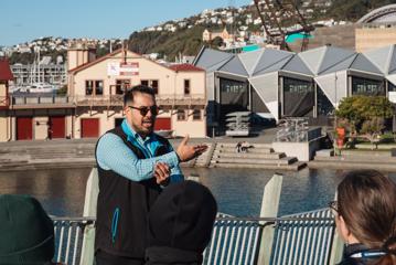 A staff member from Te Wharewaka ō Pōneke explains to a group the history of the building, land, and waka (canoe). In the background, is the Wharewaka building and The Rowing CLub.