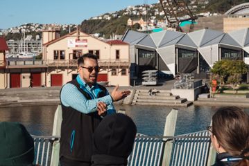 A staff member from Te Wharewaka ō Pōneke explains to a group the history of the building, land, and waka (canoe). In the background, is the Wharewaka building and The Rowing CLub.
