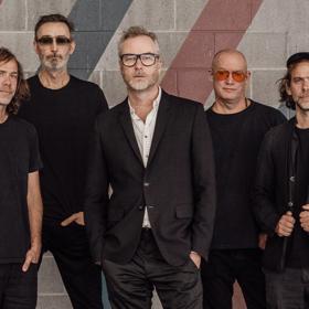 The five band members of The National stand in front of a grey brick wall wearing black with serious facial expressions. 