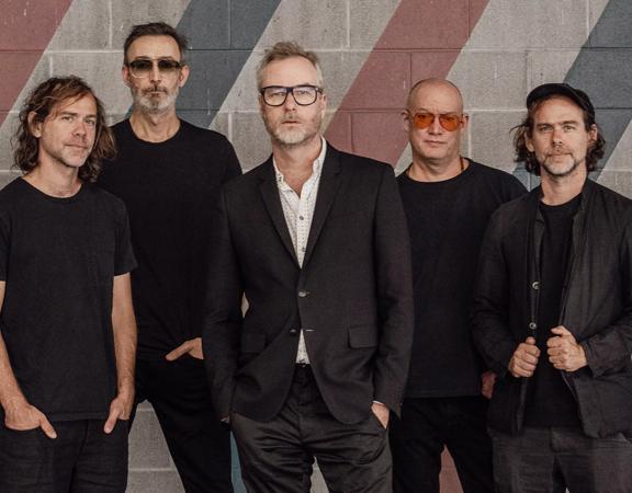 The five band members of The National stand in front of a grey brick wall wearing black with serious facial expressions. 