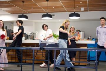 A blond young woman in a black top and light blue jeans walks and smiles with five colleagues standing in the background and a ping pong table in the foreground in a modern office kitchen.