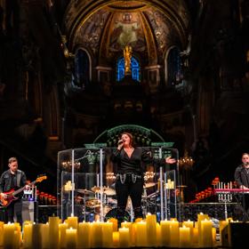 Three musicians wearing all-black perform by candlelight at a church.