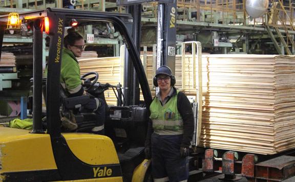 1 Juken wokrer smiles for the camera while the other drives a forklift with large amounts of wood sheets on the forks.