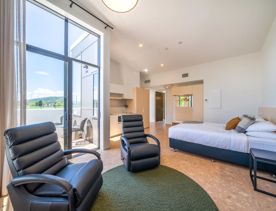 Accommodation room at NZCIS. Image from corner of the room with bed to the right, kitchenette and entrance in the background, and black armchairs and view of a balcony in the foreground.