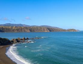 Breaker Bay on a sunny day, blue and green waves crashing on the stoney shore, with green cliffs surrounding.