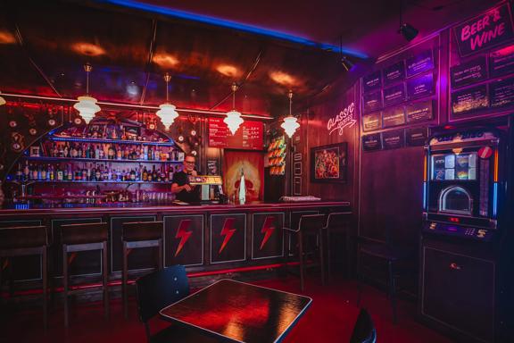 The interior of Humdinger Bar, red and blue neon lights illuminate the wood-paneled space. A bartenders is pouring a pint and there is a retro American dive-bar vibe.