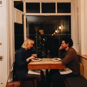A couple on a dinner date enjoy food and wine at a small table by the front window at Rita, a small restaurant in Aro Valley, Wellington.