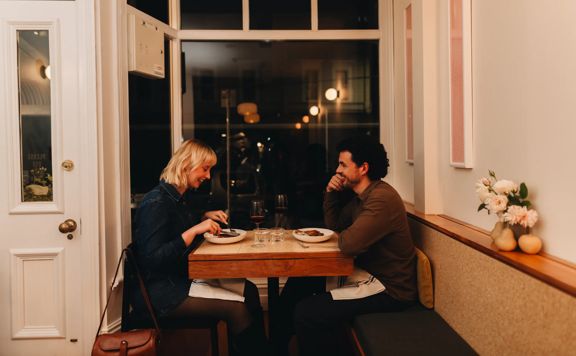 A couple on a dinner date enjoy food and wine at a small table by the front window at Rita, a small restaurant in Aro Valley, Wellington.