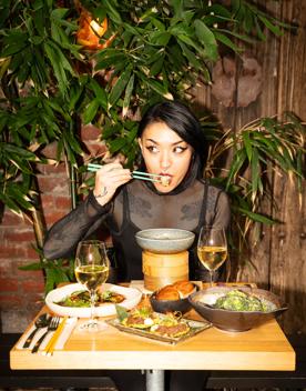 A chic-looking person dressed in all black sits at a small table with two glasses of white wine and four plates of food. They're looking to their right while using teal chopsticks to eat. 