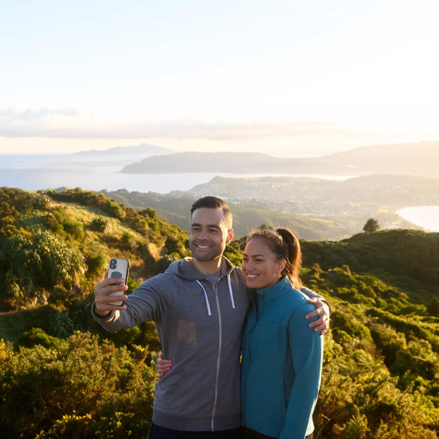 2 people posing for a selfie at the top of Mount Rangituhi, with a view of Porirua behind them.