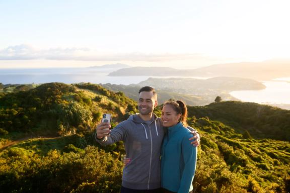 2 people posing for a selfie at the top of Mount Rangituhi, with a view of Porirua behind them.