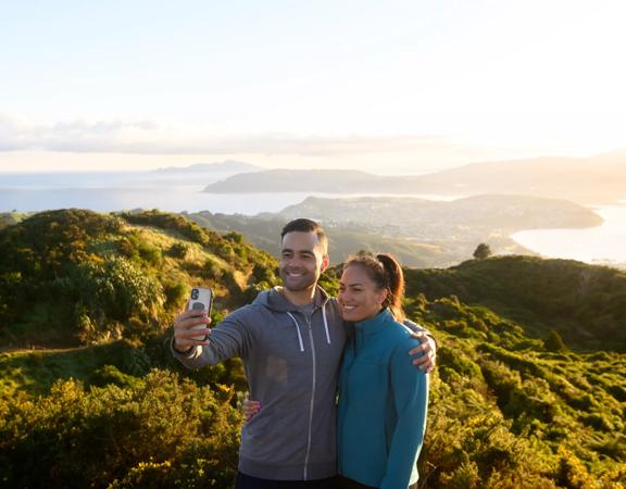 2 people posing for a selfie at the top of Rangituhi, with a view of Porirua behind them.