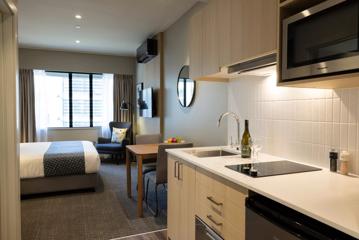 Inside a hotel suite with a small kitchenette, a table with two chairs, a queen-sized bed and an armchair in the corner by the window.