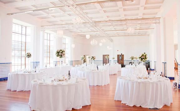 A large bright room, set up for a banquet with round tables with white tablecloths, at Massey University in Wellington, New Zealand. 