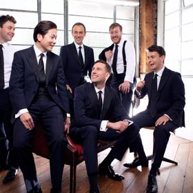 The six vocalists in The King’s Singers are all dressed in dark blue suits, some with their jackets off in an informal manner. They are sitting or standing in a light-filled room.