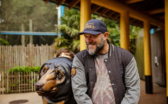 A person in a navy blue cap with "Cocaine Bear" in yellow writing smiles and leans on a brown bear statue and another person is peaking out from behind the bear.