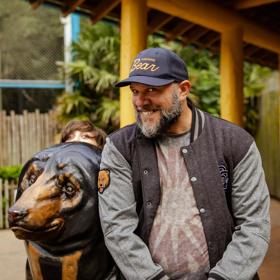 A person in a navy blue cap with "Cocaine Bear" in yellow writing smiles and leans on a brown bear statue and another person is peaking out from behind the bear. 