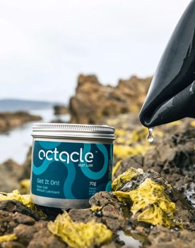 A tin of Octacle wetsuit lube on a rock with a wetsuit dripping water on the right.
