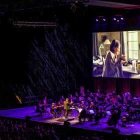 A full orchestra performs under purple floodlights on stage, with the film Love Actually playing on a projector above them.