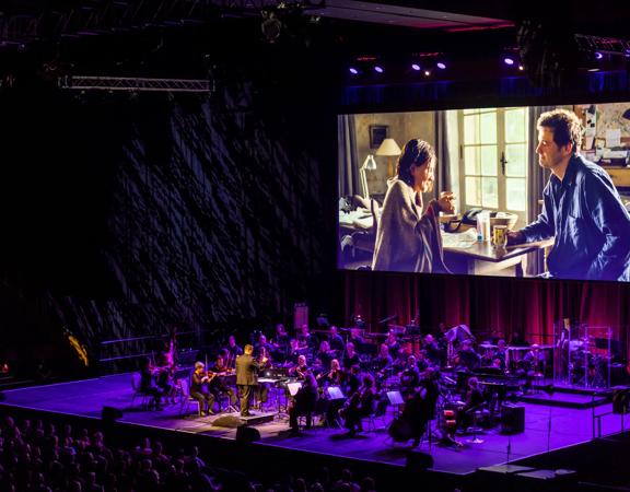 A full orchestra performs under purple floodlights on stage, with the film Love Actually playing on a projector above them.