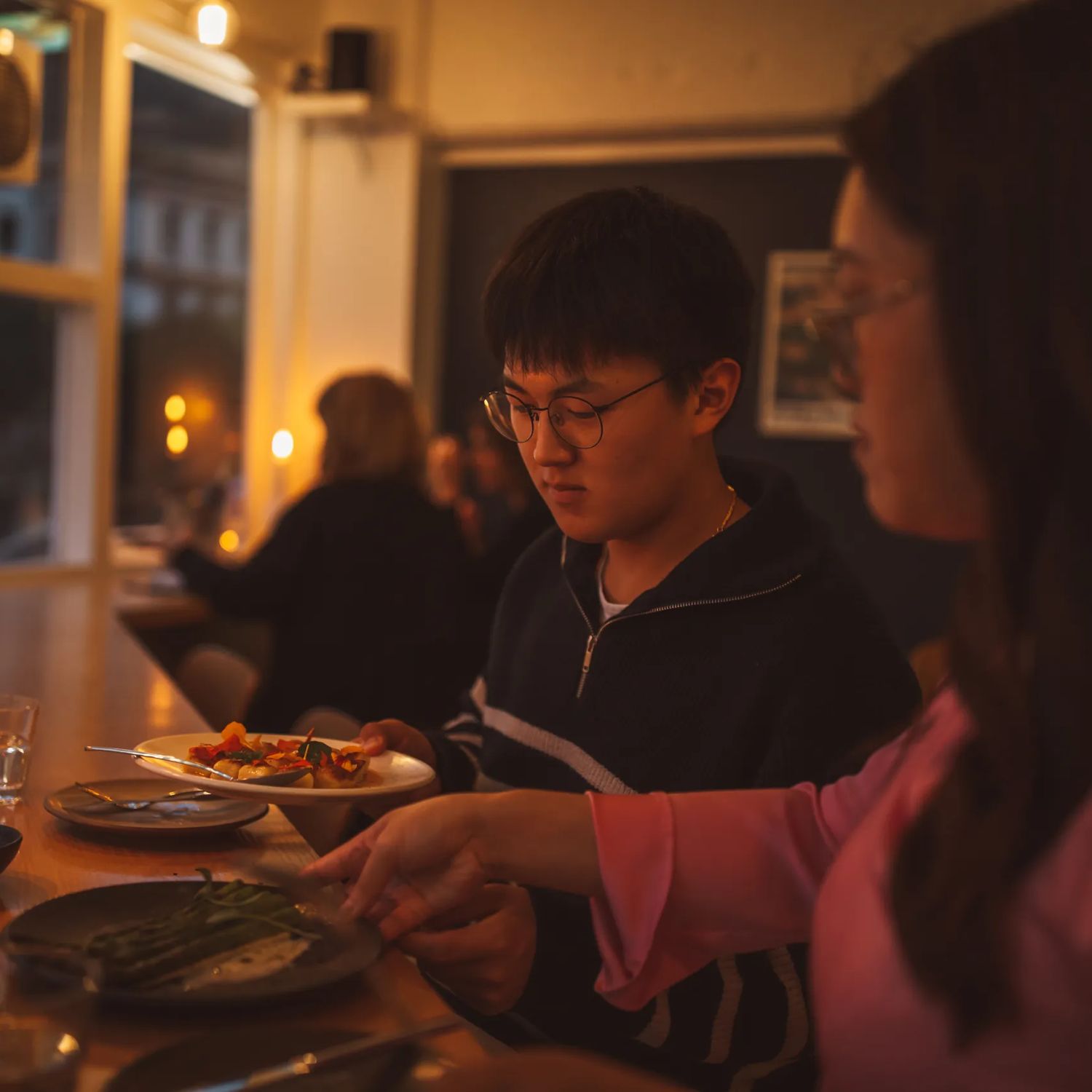 Two friends swapping plates of food inside Supra on Dixon Street. The candlelit room is dim, with people enjoying their meals in the background.