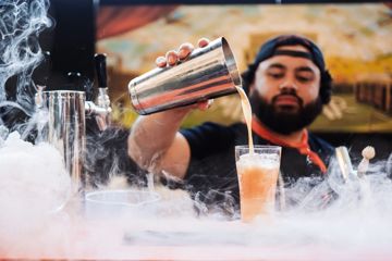 A bartender pours a drink from a metal shaker into a pint glass with smoke covering the countertop.