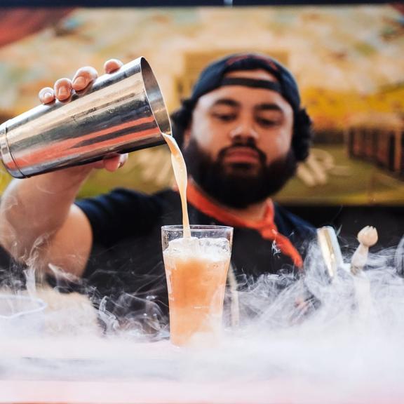A bartender pours a drink from a metal shaker into a pint glass with smoke covering the countertop.
