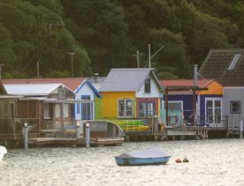 The Mana Boatsheds in Porirua, New Zealand. Two boats are floating in front and a green lush forest is seen behind the houses.