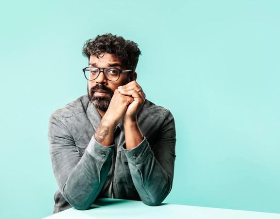 British comedian, presenter, and actor, Jonathan Romesh Ranganathan, poses wearing a grey jacket, with clasped hands and elbows resting on the edge of a table in front of a light blue wall. 