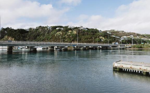 A cement bridge over a body of water with houses and hilltops visible in the background.