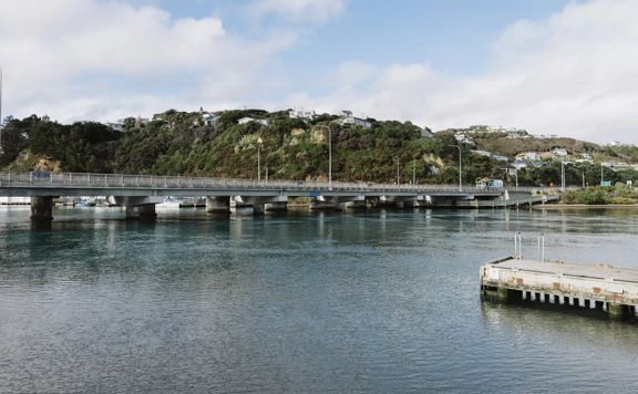 A cement bridge over a body of water with houses and hilltops visible in the background.