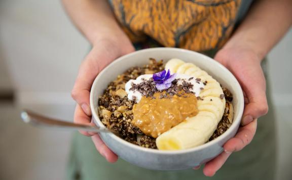 Hands holding a bowl at The Oatery, with slices banana, granola and peanut but toppings.